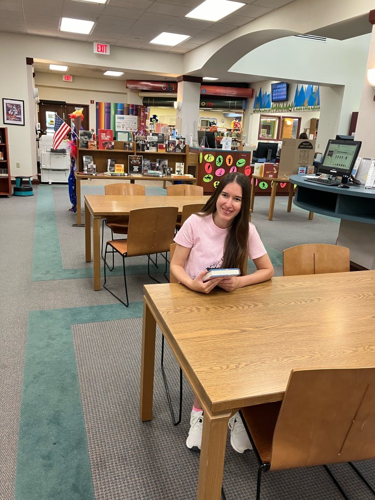Demi sitting at a table in the Murrysville Community Library and wearing her Wild Ink editor shirt